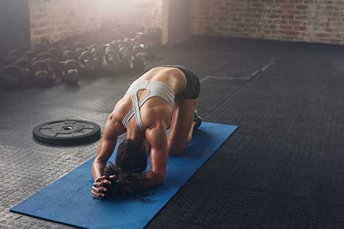 Mujer realizando ejercicios de estiramiento en una colchoneta en el gimnasio, con pesas y un disco de peso en el fondo.